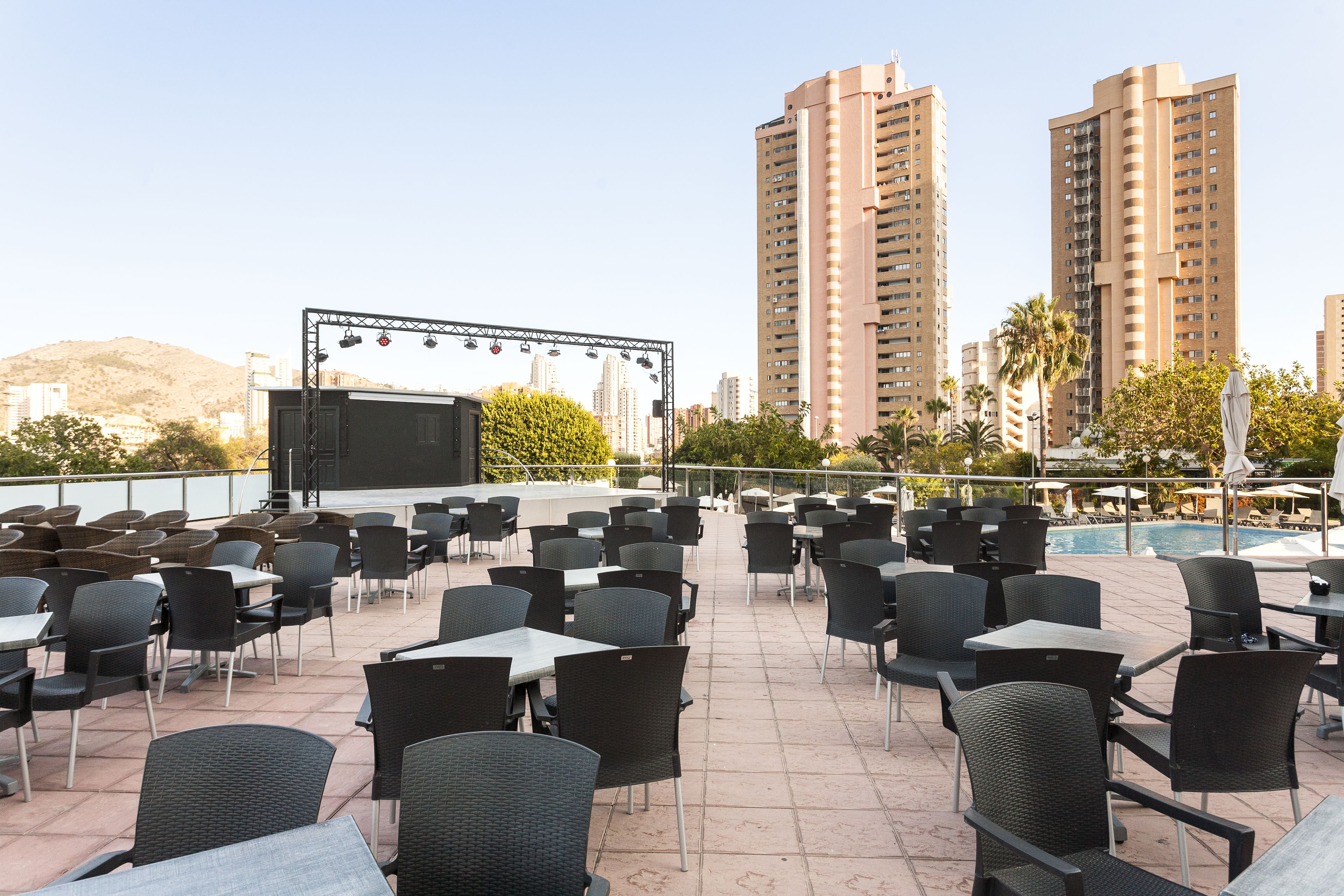 An outdoor seating area with tables and chairs, a stage with lighting, and tall buildings in the background.
