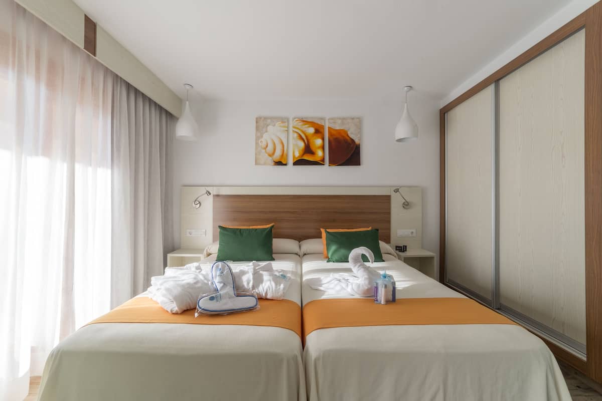 Une chambre d’hôtel avec deux lits, une tête de lit en bois, une grande fenêtre avec des rideaux légers et des œuvres d’art au-dessus des lits.