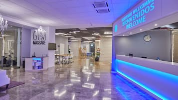 A modern reception area with a 'GTVI MASSAGE' sign, a reception desk, and a 'BIENVENIDOS WELCOME' sign.