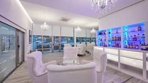 Un bar moderno con mobiliario blanco, candelabros y una barra bien iluminada repleta de botellas.