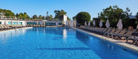 A clear blue pool with lounge chairs and umbrellas, surrounded by trees and buildings.