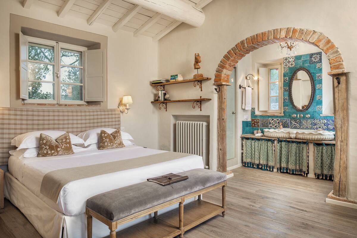 Un dormitorio con una cama grande, un banco, una ventana y un baño con pared de azulejos y un espejo.