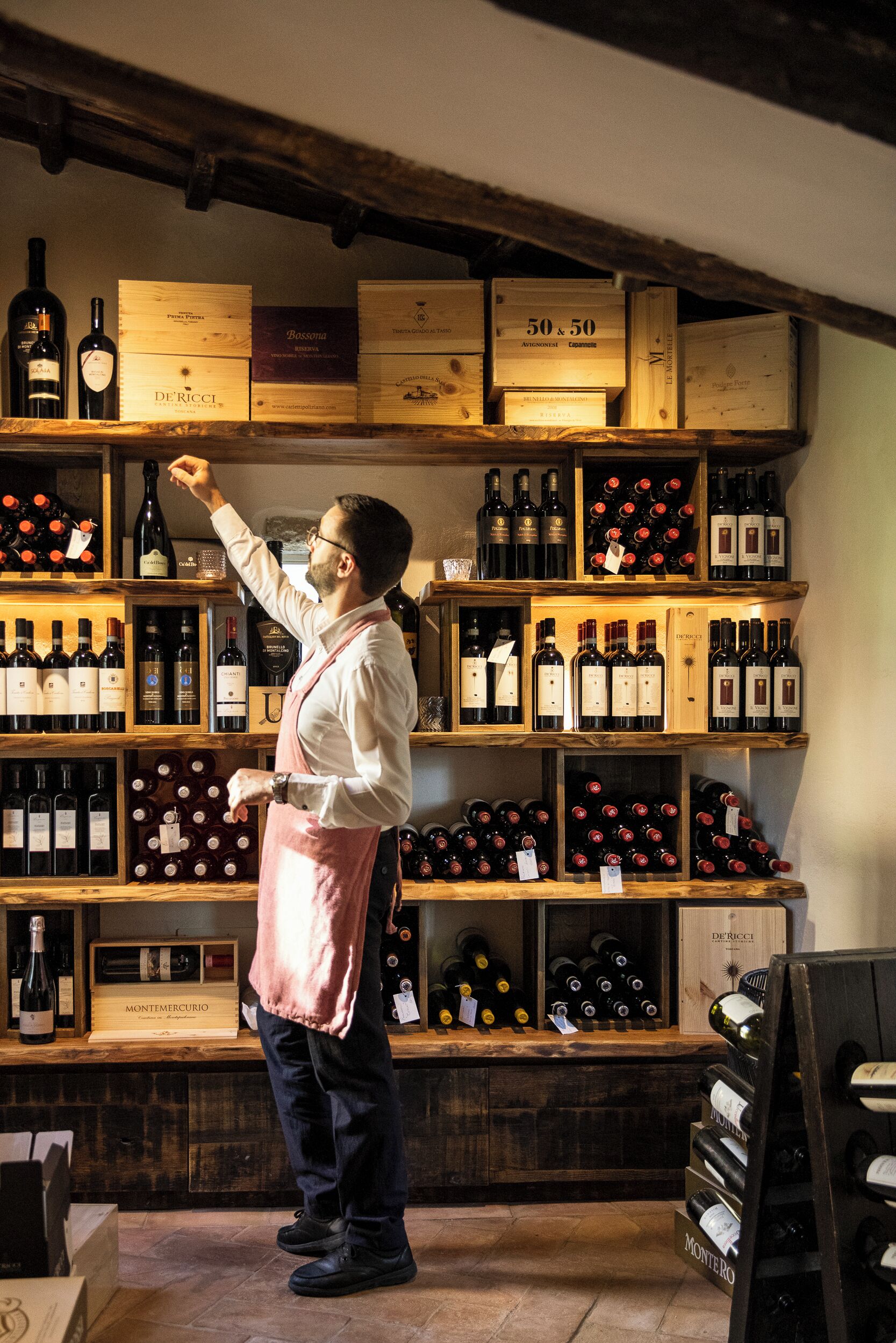 A wine cellar with wooden shelves stocked with wine bottles and boxes labeled with names like "DE RICCI" and "50 x 50."