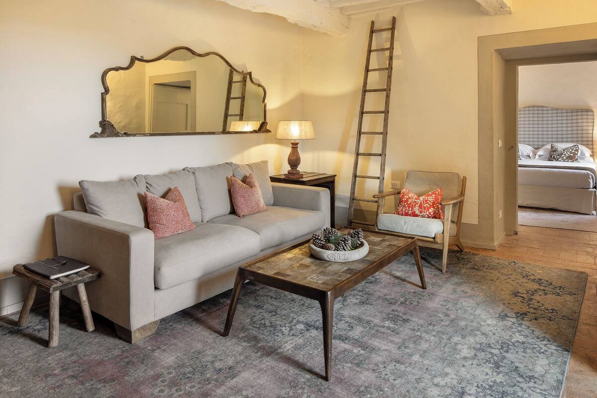 Una sala de estar con un sofá, una mesa de centro, una mesita auxiliar y una silla. Una escalera está apoyada contra la pared, y se ve una cama al fondo.