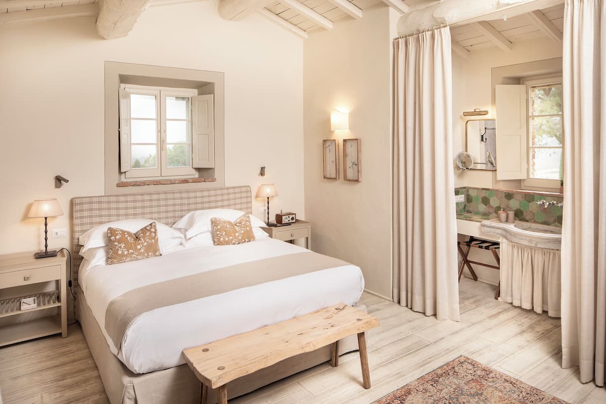 Un dormitorio con una cama grande, mesitas de noche, un banco, una ventana con cortinas, un espejo y un baño con lavabo y espejo.