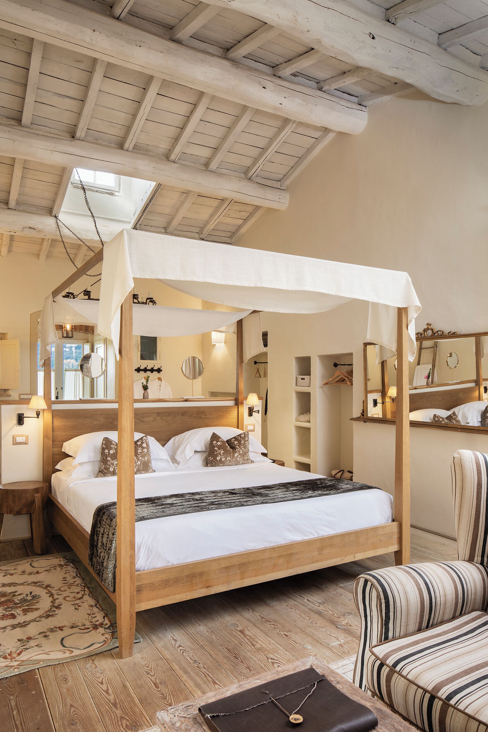 Un dormitorio con cama con dosel, vigas de madera en el techo, un espejo grande y un sillón a rayas.
