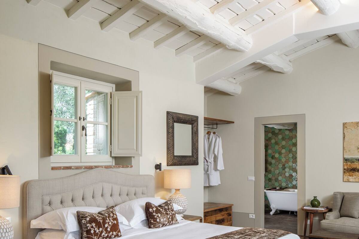 Un dormitorio con una cama grande, un banco de madera, una mesita de noche, un aparador y un baño visible a través de una puerta abierta.