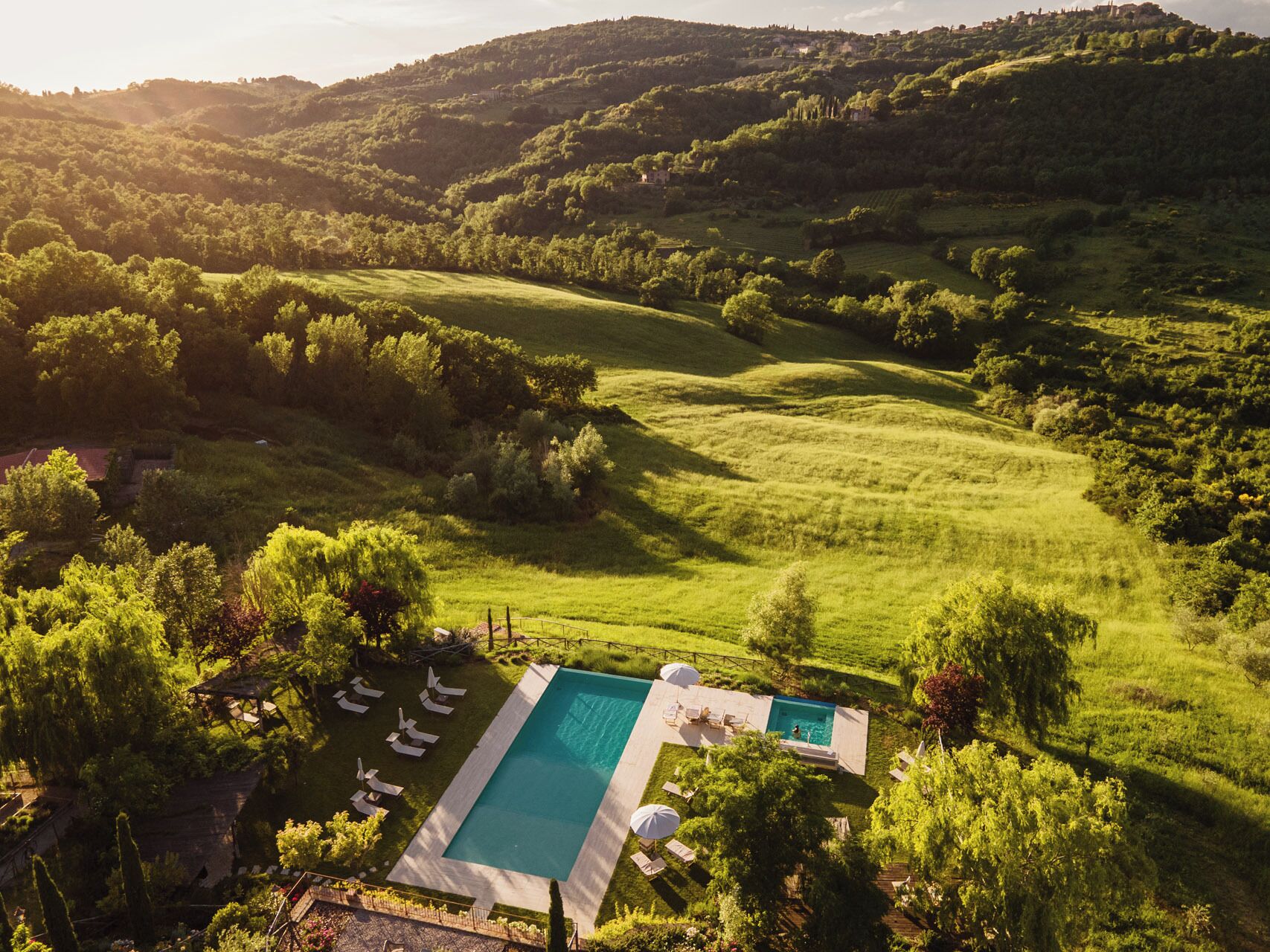 Eine Vogelperspektive auf eine üppige grüne Landschaft mit sanften Hügeln, einem Swimmingpool und einer Terrassenanlage mit Liegestühlen und Sonnenschirmen.