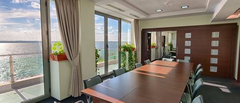 A conference room with a large table and chairs, large windows with a view of the sea, and a door leading to another room.