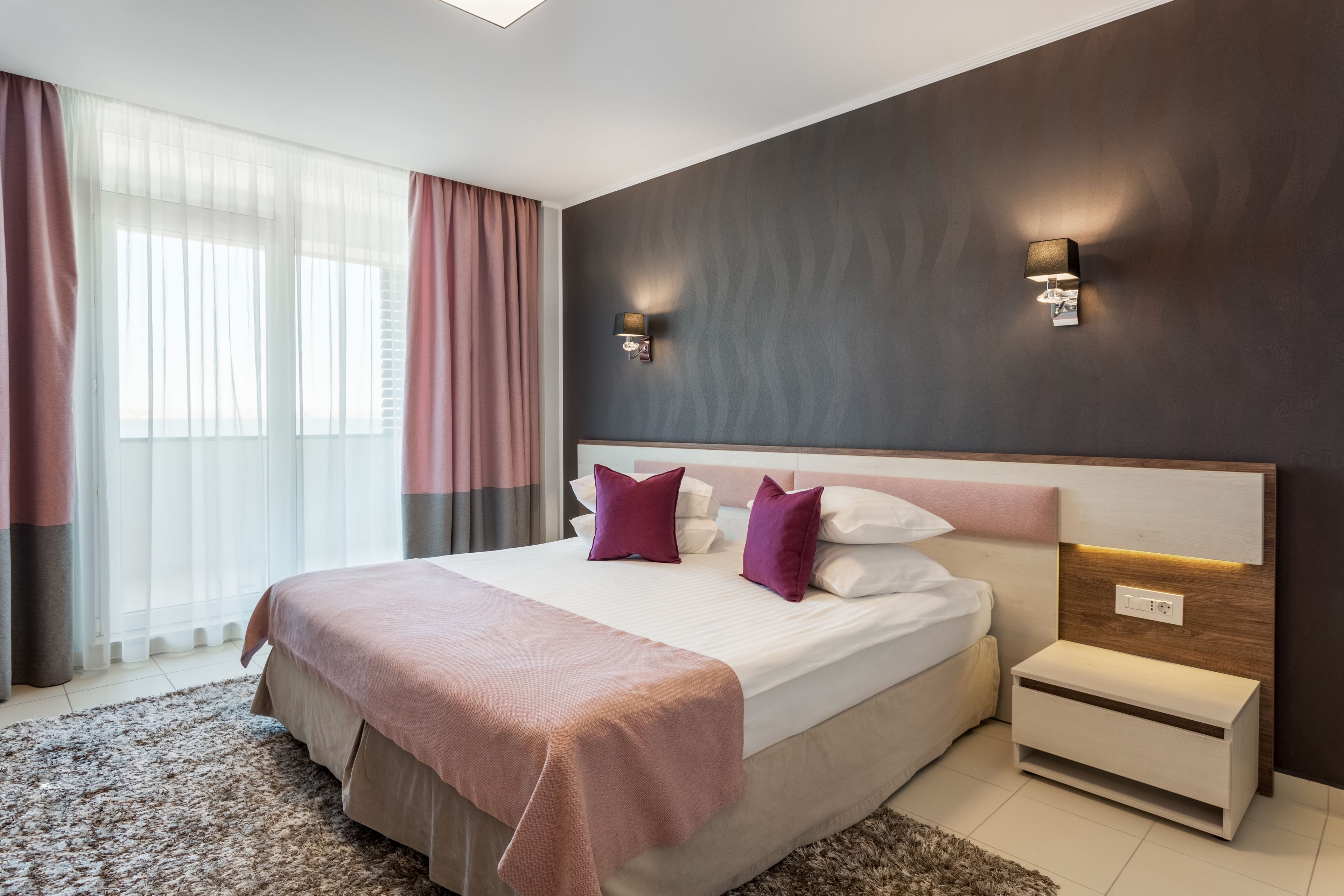 A modern bedroom with a large bed, two bedside tables, a nightstand, and a wall-mounted lamp.