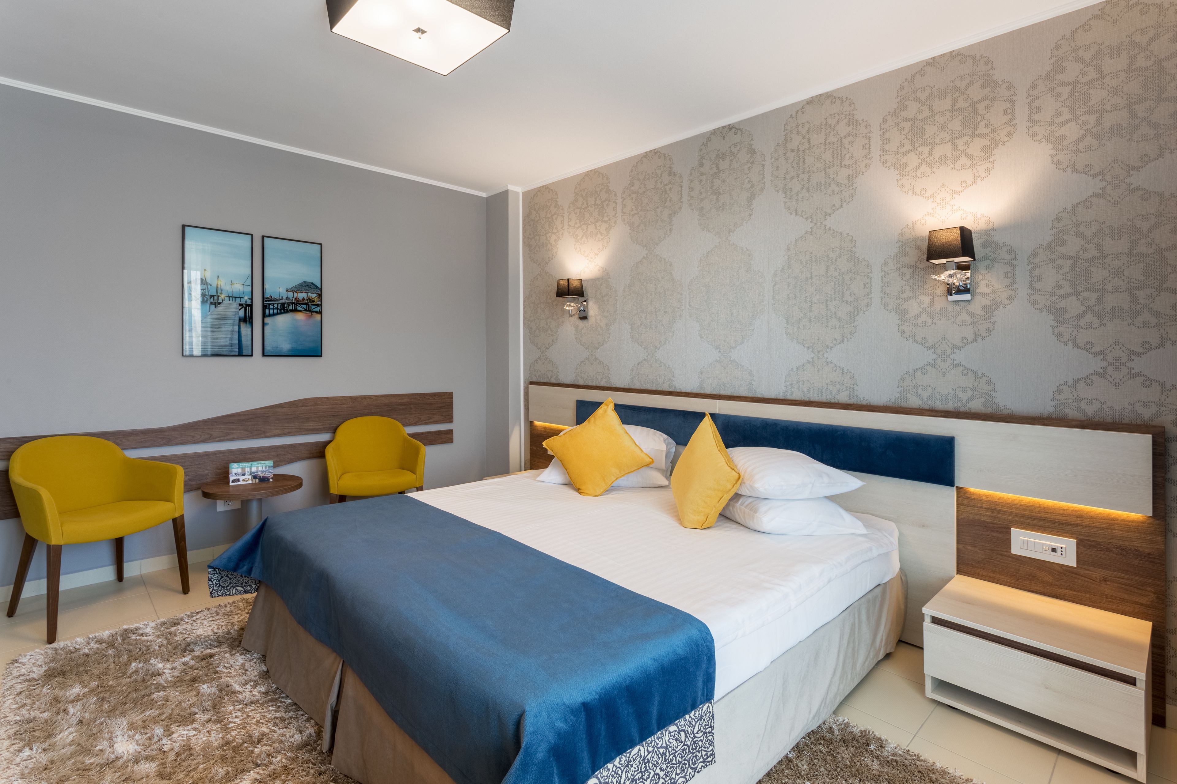 A modern hotel room with a large bed, two yellow chairs, a small table, and a nightstand.