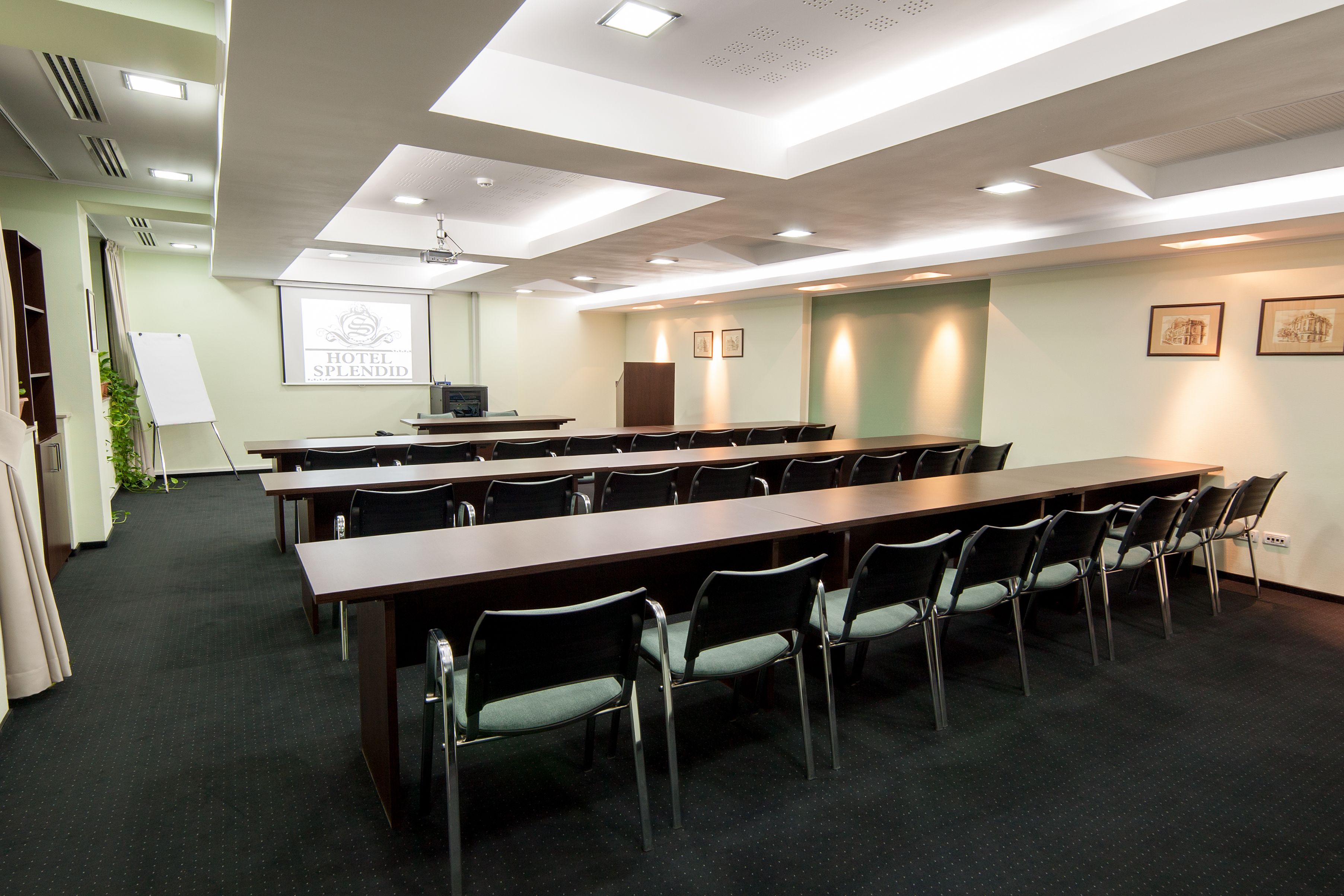 Una sala conferenze con un lungo tavolo, sedie, una lavagna e uno schermo proiettore che mostra "HOTEL SPLENDID".