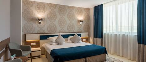 A modern hotel room with a large bed, two bedside tables, a nightstand, a sofa, and a view of the city through sheer curtains.