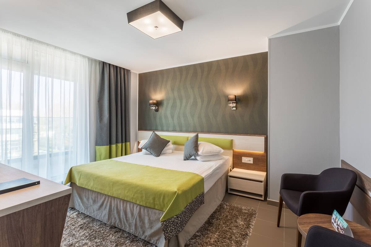 A modern hotel room with a large bed, a desk, a chair, and a nightstand.