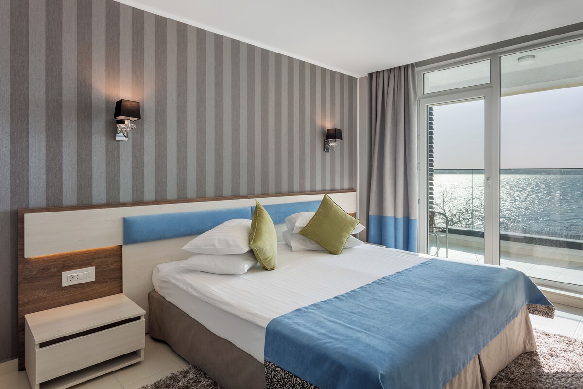 A modern hotel room with a large bed, a nightstand, a wall-mounted lamp, and a view of the sea through a large window.