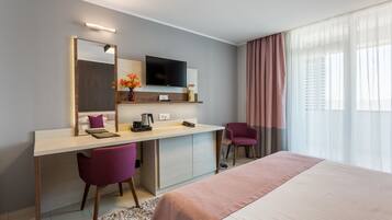 A modern hotel room with a bed, a desk with a chair, a television, and a window with curtains.