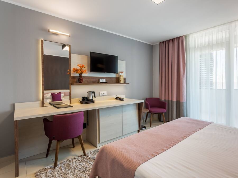 A modern hotel room with a bed, a desk with a chair, a television, and a window with curtains.
