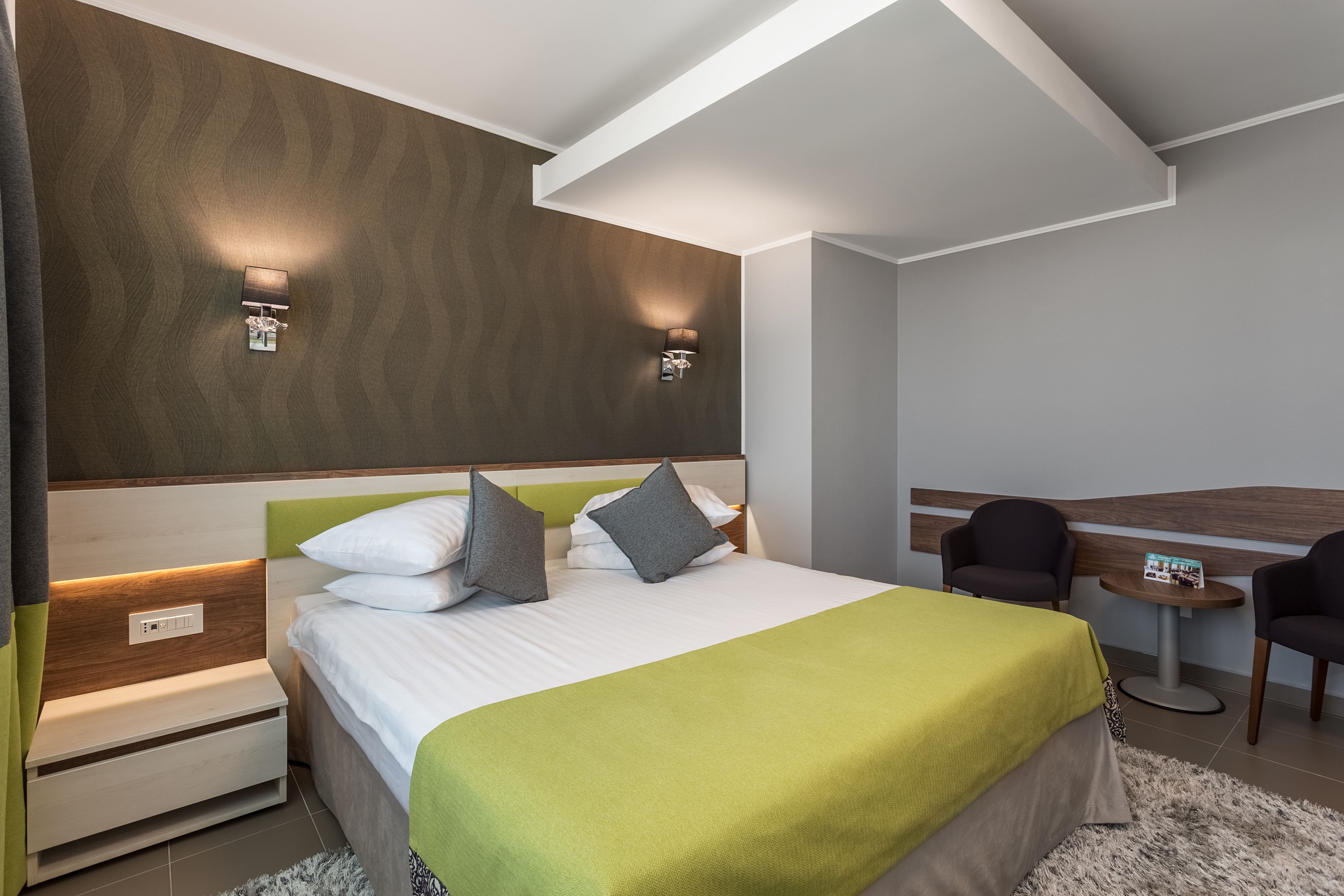 Een moderne hotelkamer met een opgemaakt bed, twee bedlampen, een tafeltje met een stoel en een hoofdbord met ingebouwde verlichting.