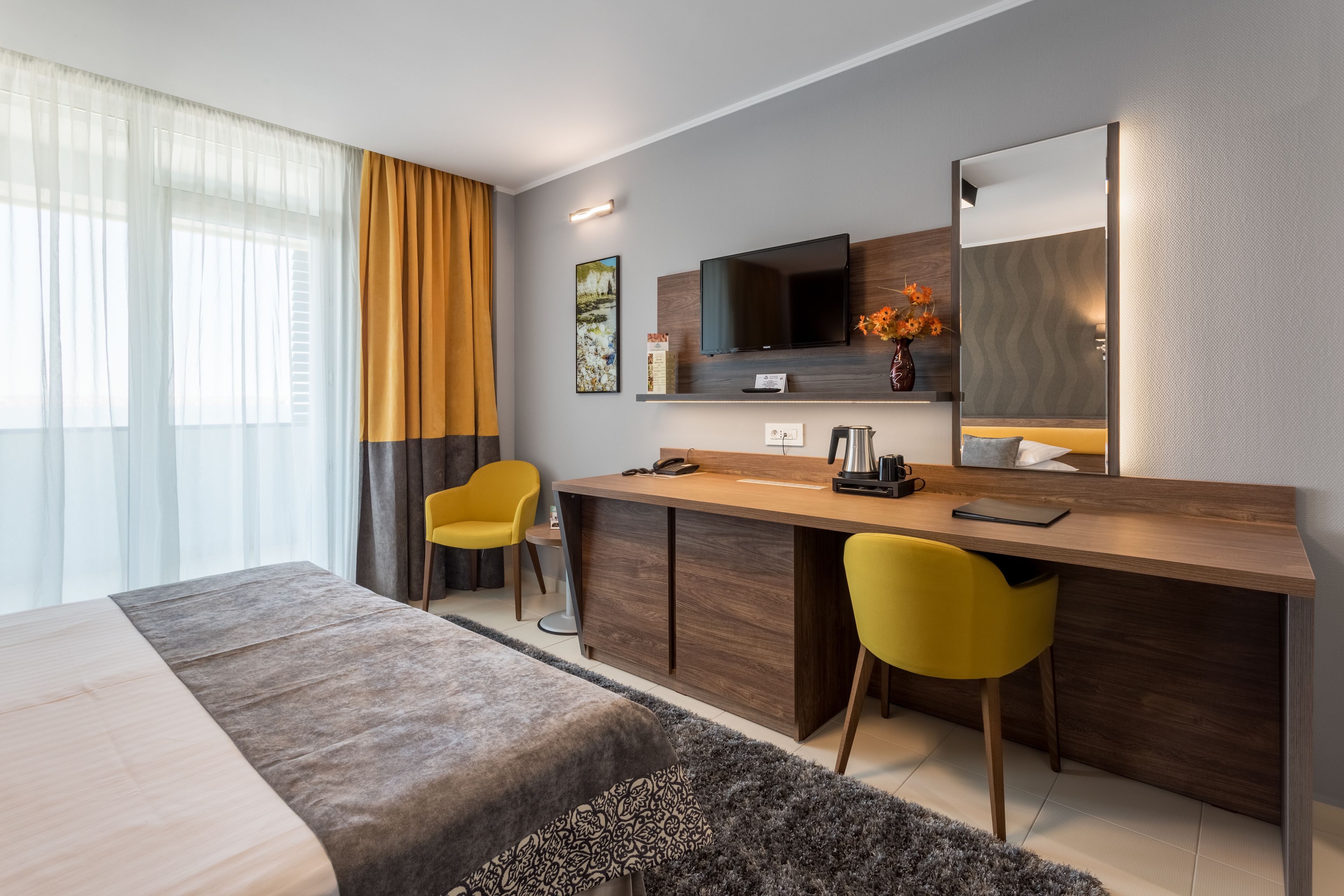 Habitación de hotel moderna con cama, escritorio, dos sillas, televisión y un ventanal con cortinas.