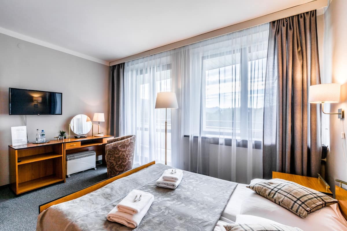 Habitación de hotel con cama, escritorio, televisor, lámpara y vistas al exterior a través de amplias ventanas.