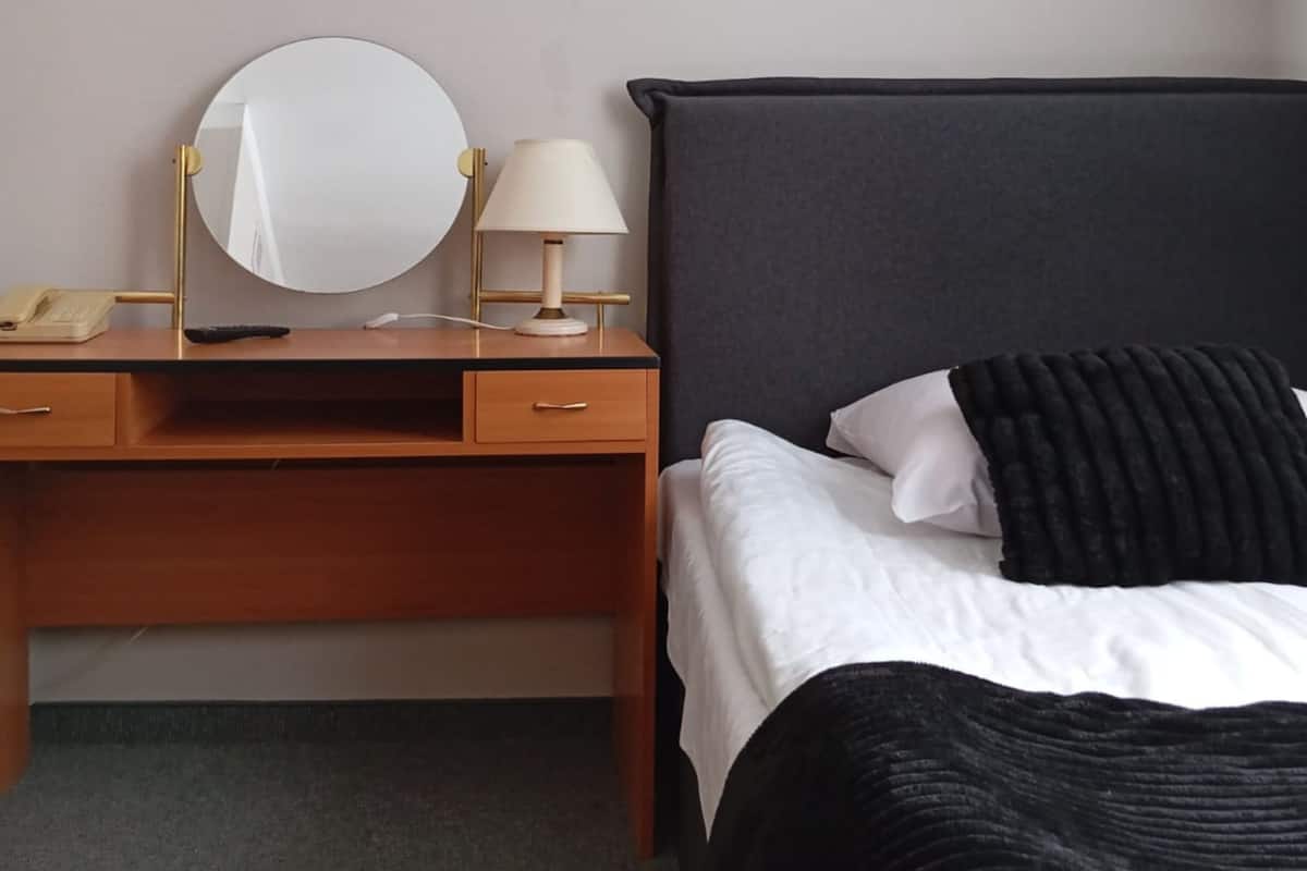 Habitación de hotel con un escritorio de madera, un espejo redondo, una lámpara, una cama con ropa de cama blanca y negra, y un televisor montado en la pared.