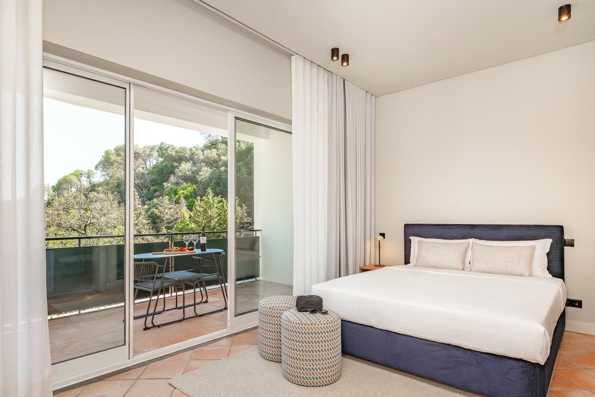 Un dormitorio con una cama grande, un balcón con mesa y sillas, y vista a árboles.