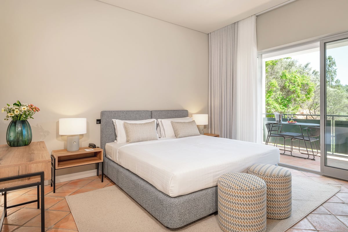 Habitación de hotel moderna con una cama grande, dos mesitas de noche, un escritorio pequeño y un balcón con vistas a árboles.