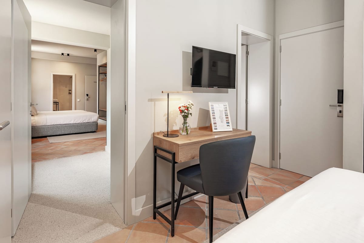 Habitación de hotel con cama, escritorio con silla, televisor montado en la pared y puerta que da a otra habitación.