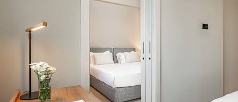 Een hotelkamer met een houten nachtkastje, een bloemenvaas, een nachttafeltje met een wit kussen en een bed met witte lakens.