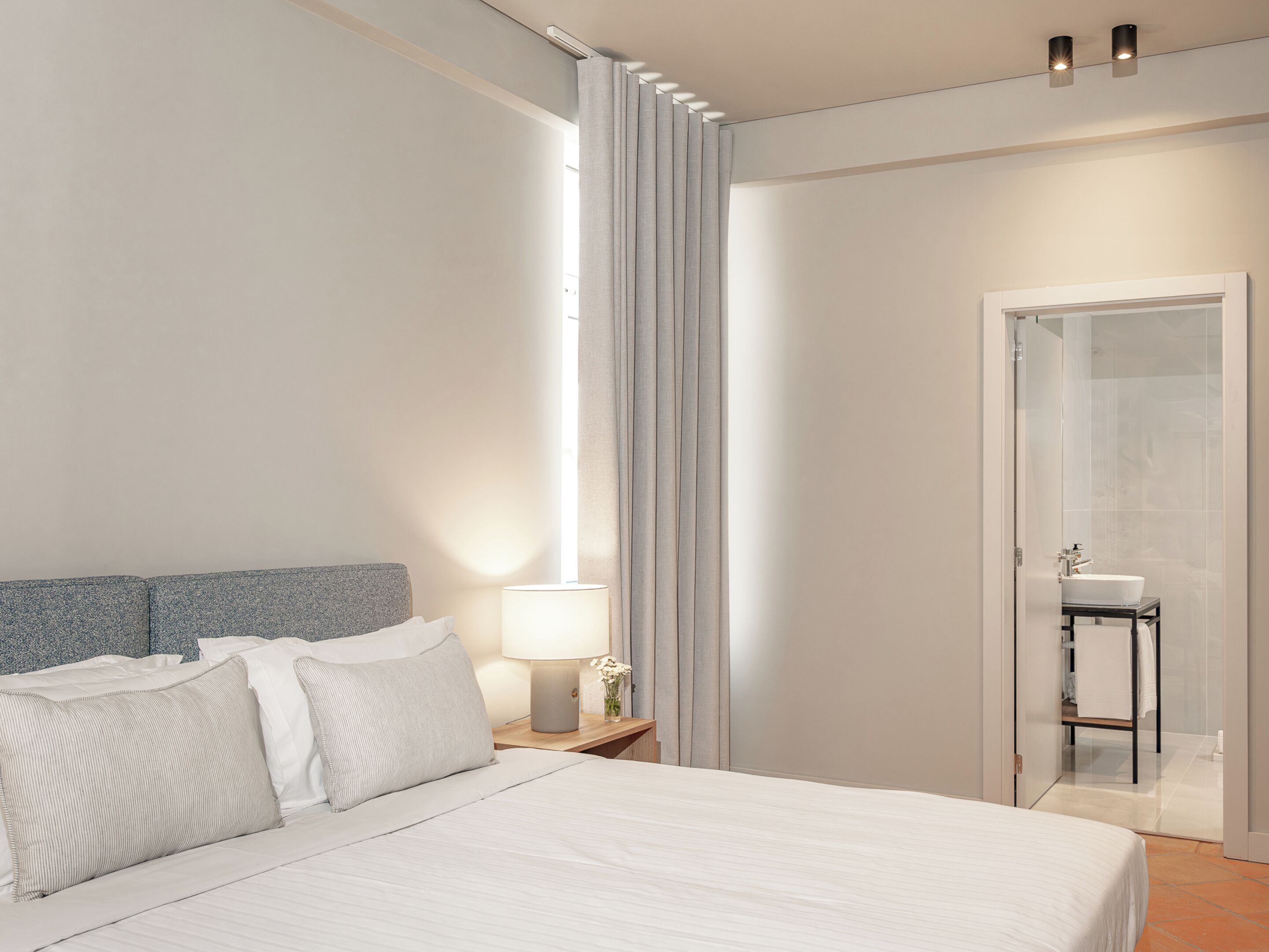 Ein modernes Hotelzimmer mit einem großen Bett, einem Nachttisch mit Lampe, einem Fenster mit Vorhängen und einem durchgegangene Tür erkennbaren Badezimmer.
