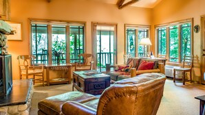 A cozy living room with large windows, a leather sofa, a wooden table, and a fireplace.
