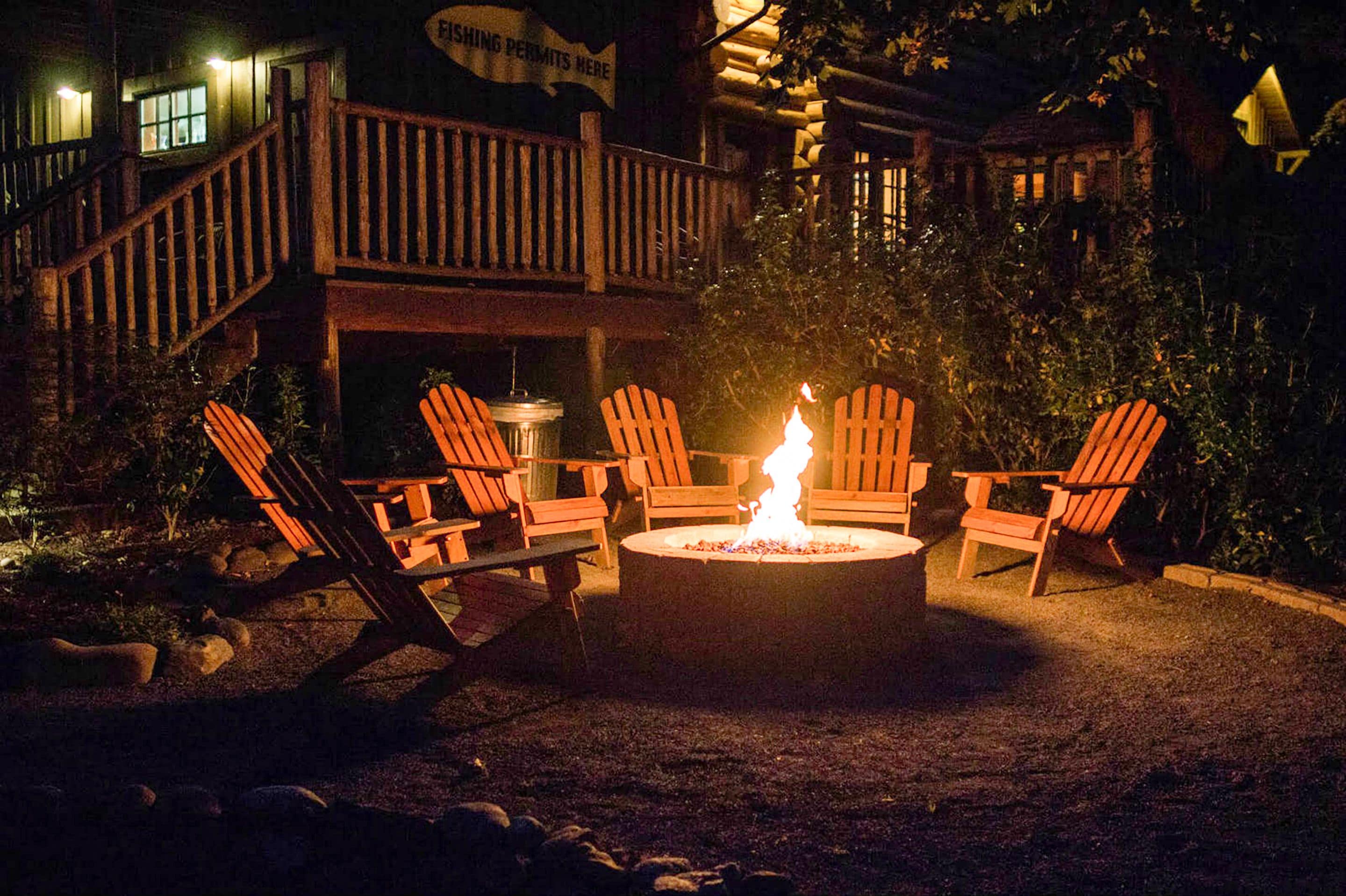 A circular fire pit with four Adirondack chairs around it, set in a gravel area with a wooden deck and stairs leading to a building with a sign that reads "FISHING PERMITS HERE".