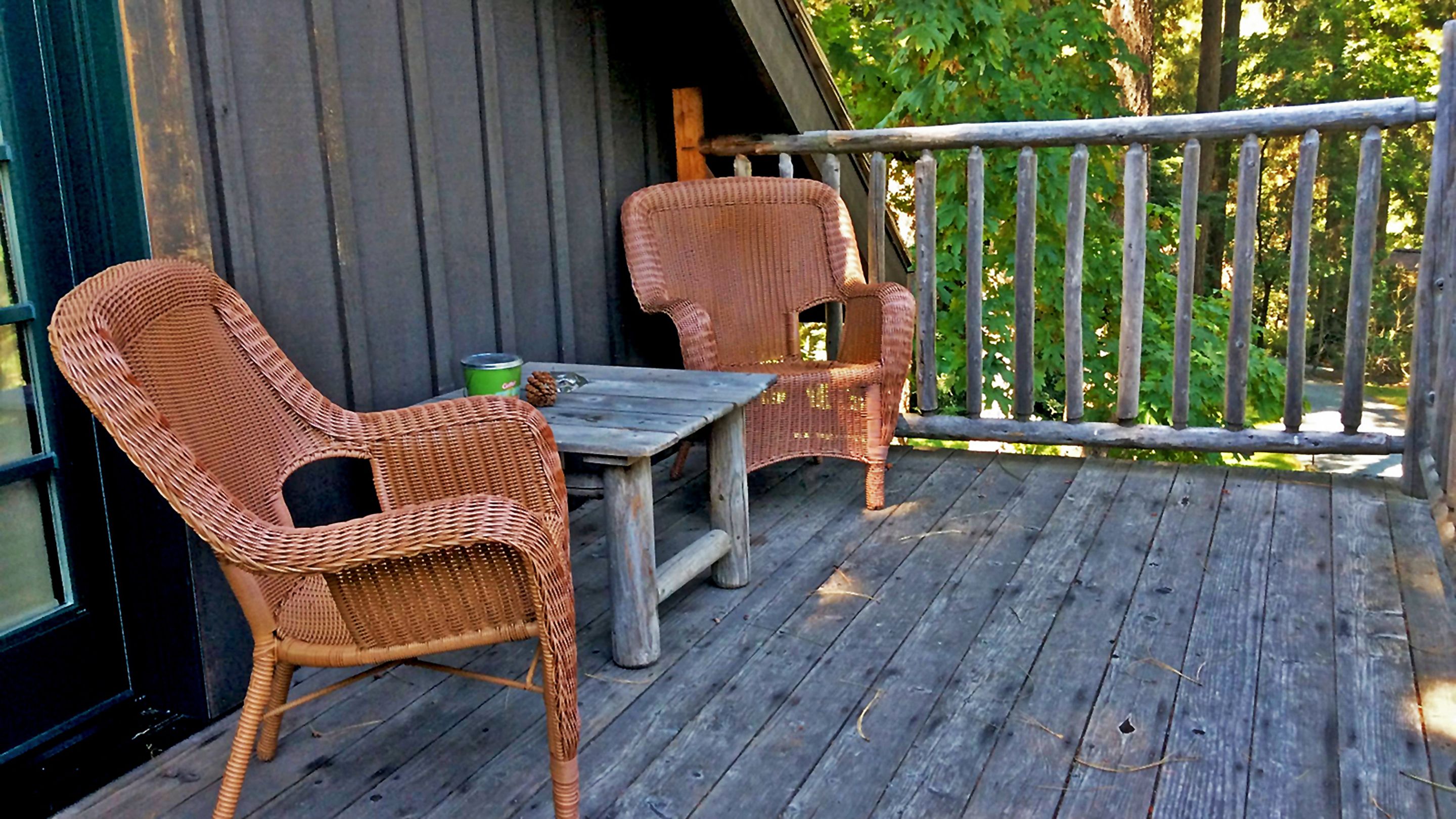 Une terrasse en bois avec des chaises en osier et une table, une canette de Coca-Cola et une pomme de pin posée sur la table.