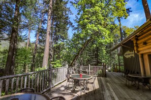 A wooden deck with outdoor seating, surrounded by tall trees and a clear sky.