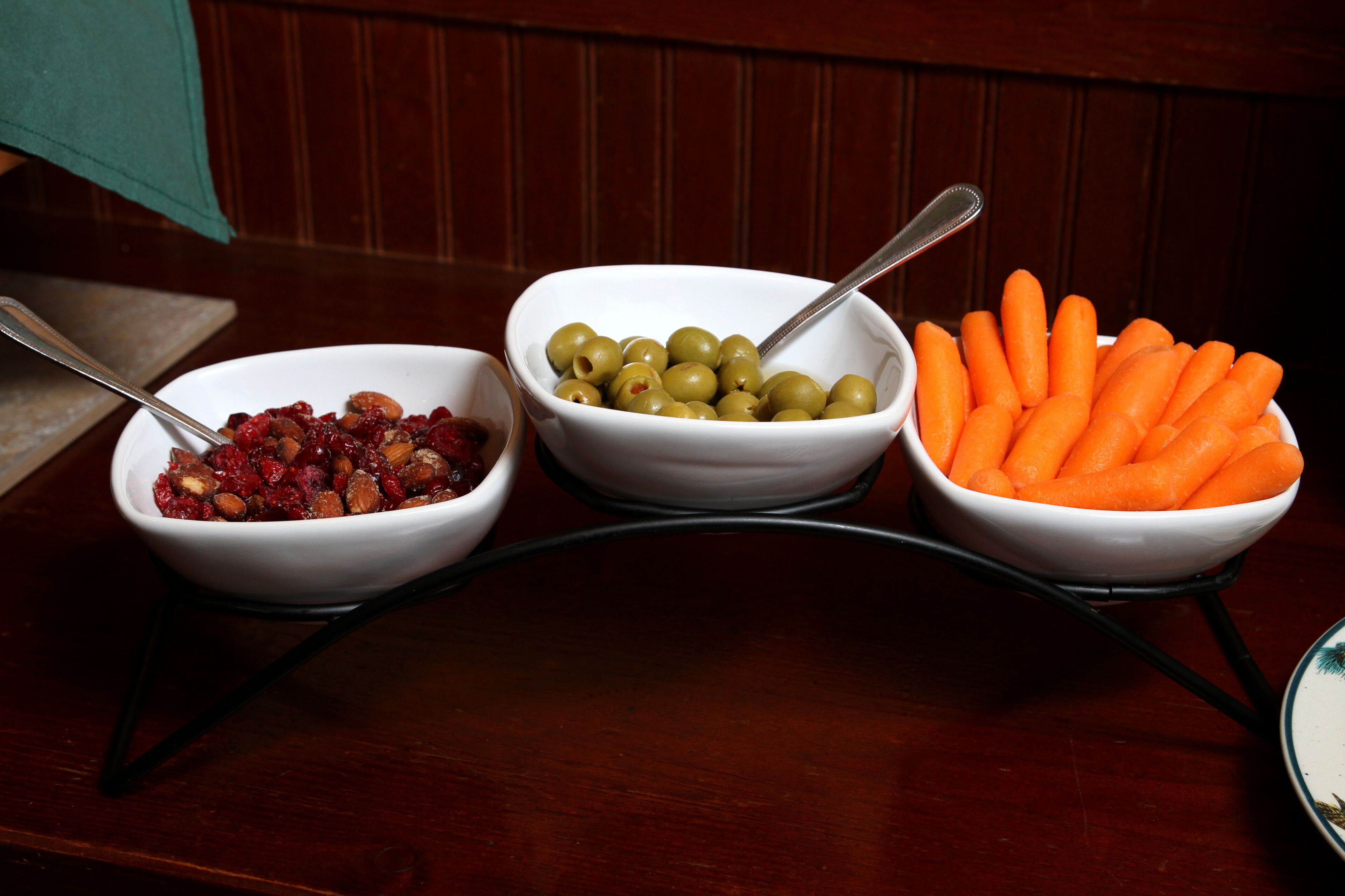 Three white bowls on a black stand, each containing different food items: dried fruits, green olives, and carrots.