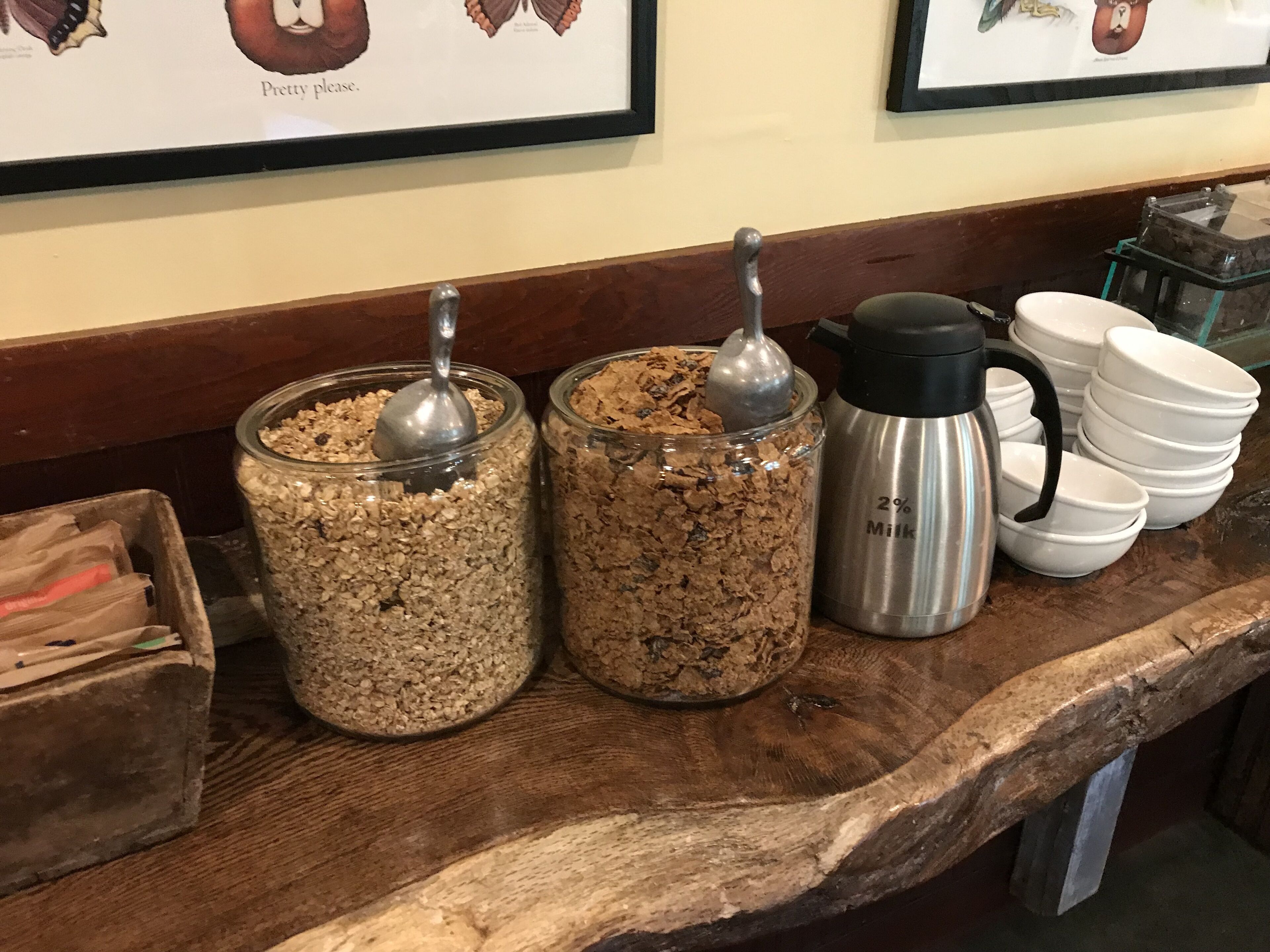 Un bar de petit-déjeuner avec deux bocaux en verre remplis de granola, un récipient à lait en acier inoxydable et des bols blancs empilés.