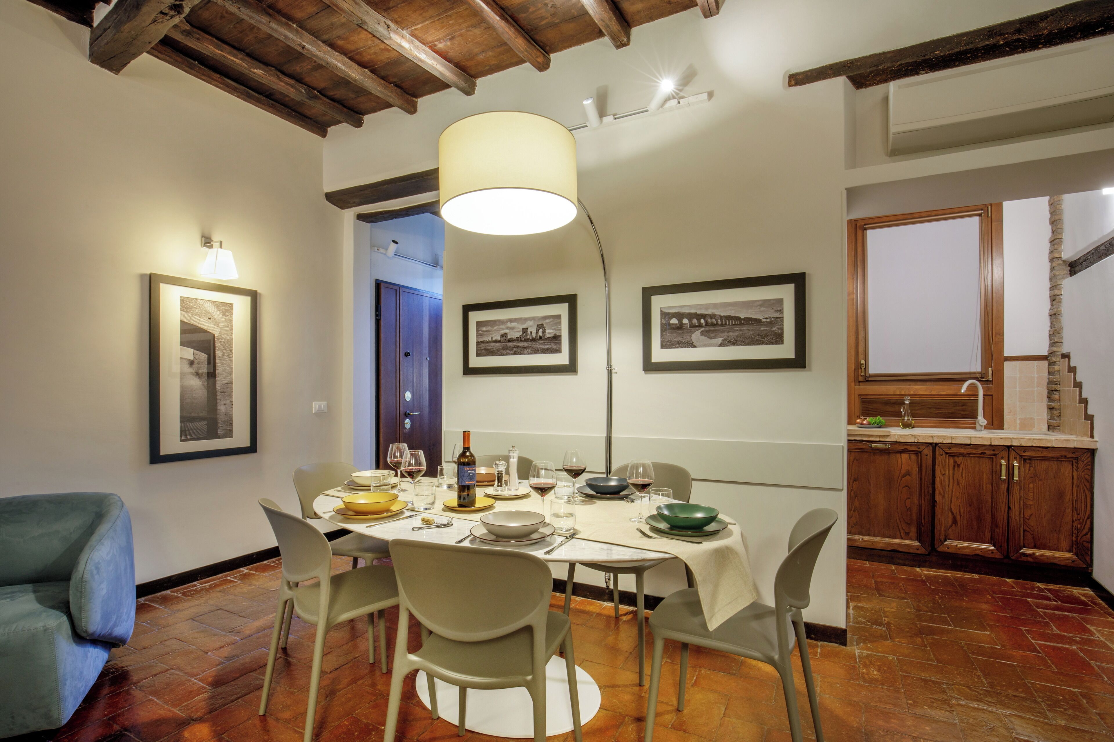 A dining area with a round table, chairs, and a chandelier. There are framed pictures on the wall and a staircase leading to another room.