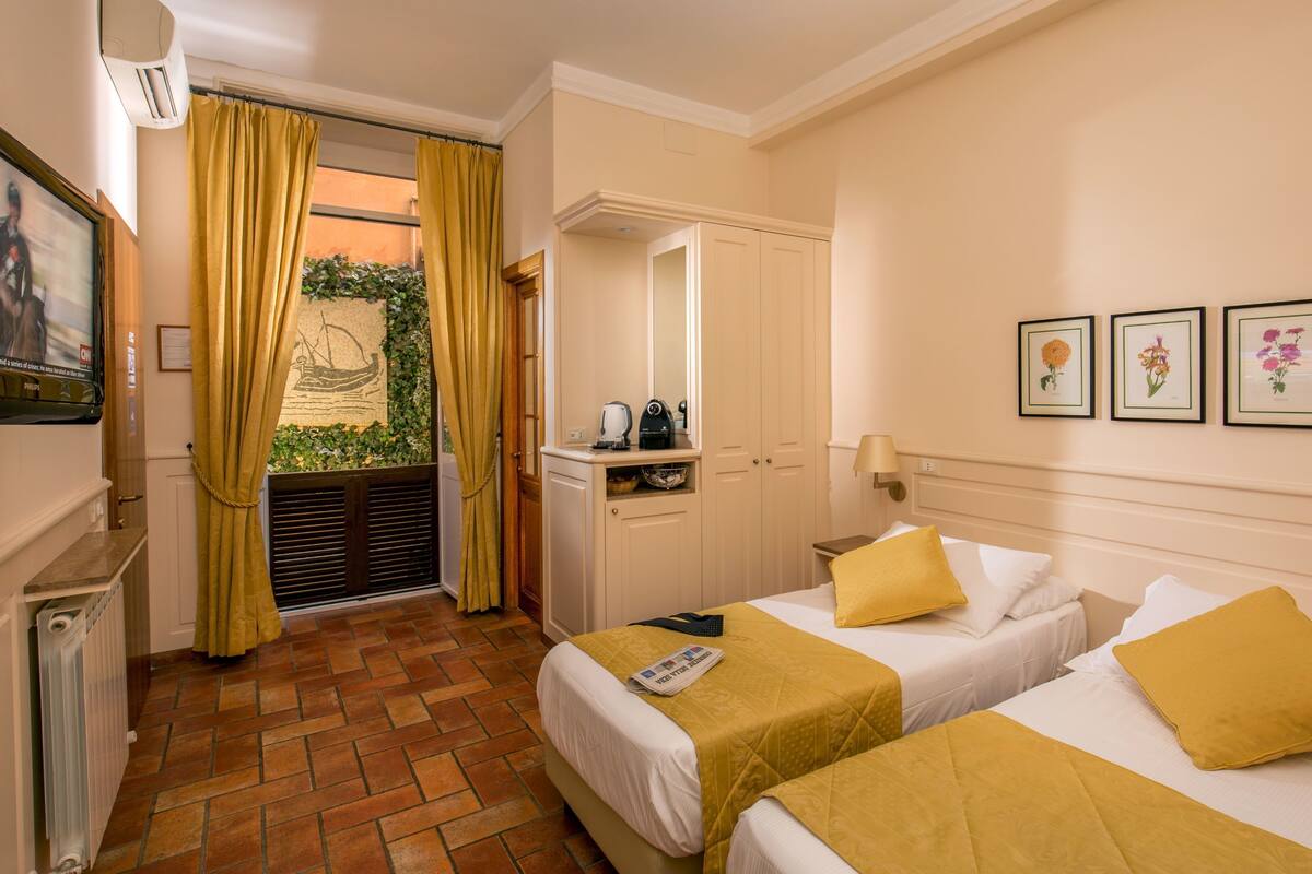 A hotel room with two beds, a television, a desk with a kettle, and a window with curtains.