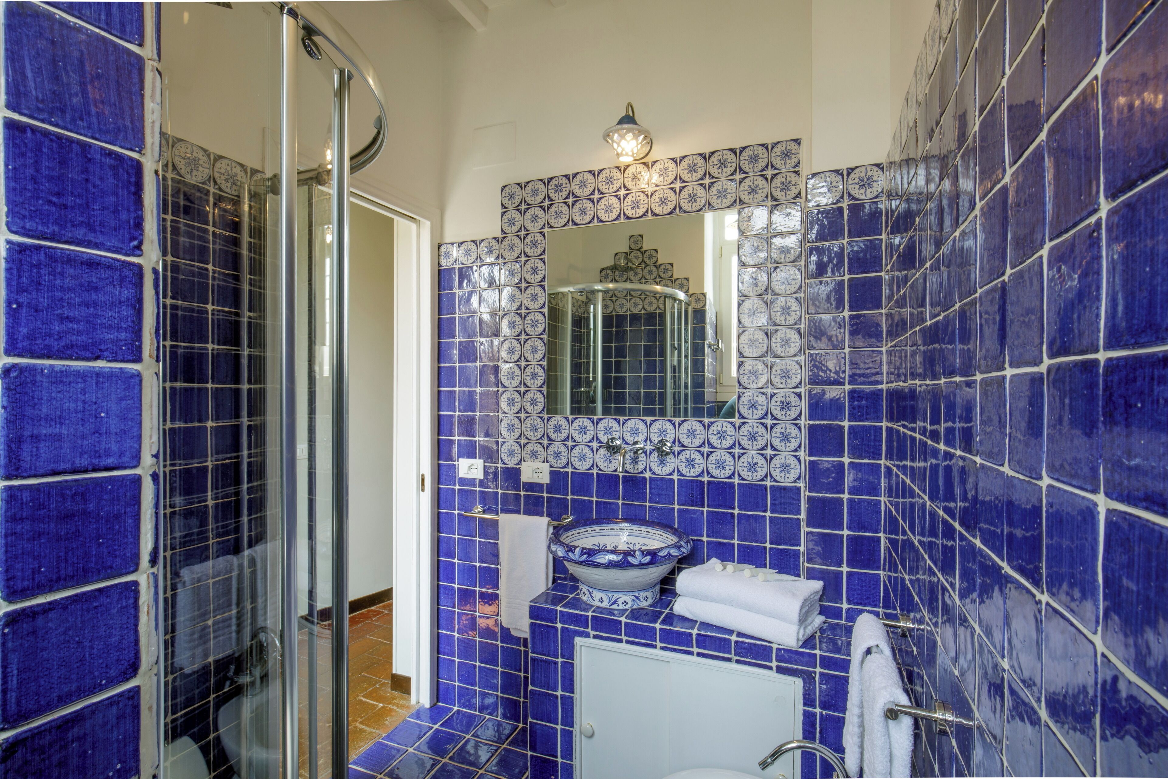 Un baño con azulejos azules, cabina de ducha con mampara de vidrio, lavabo con pileta decorativa y espejo sobre el lavabo.