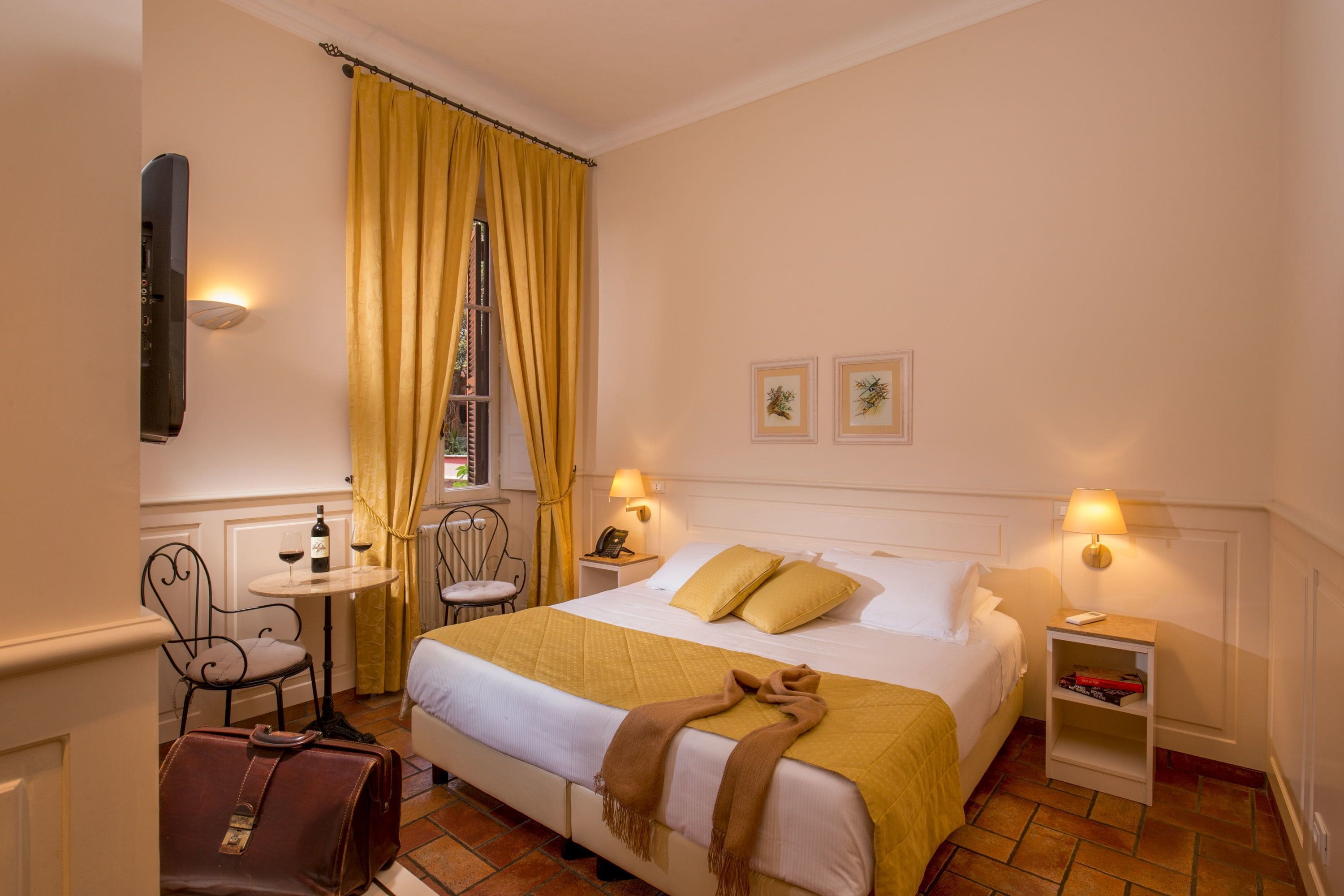 A hotel room with a bed, bedside tables, a chair, a small table with a bottle of wine, and a suitcase.