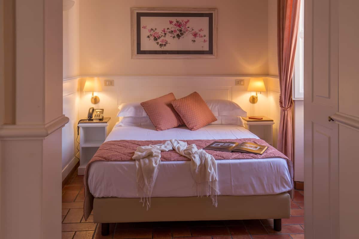 A neatly made bed with a pink blanket and pillows, bedside lamps, a framed floral artwork on the wall, and a book on the bed.