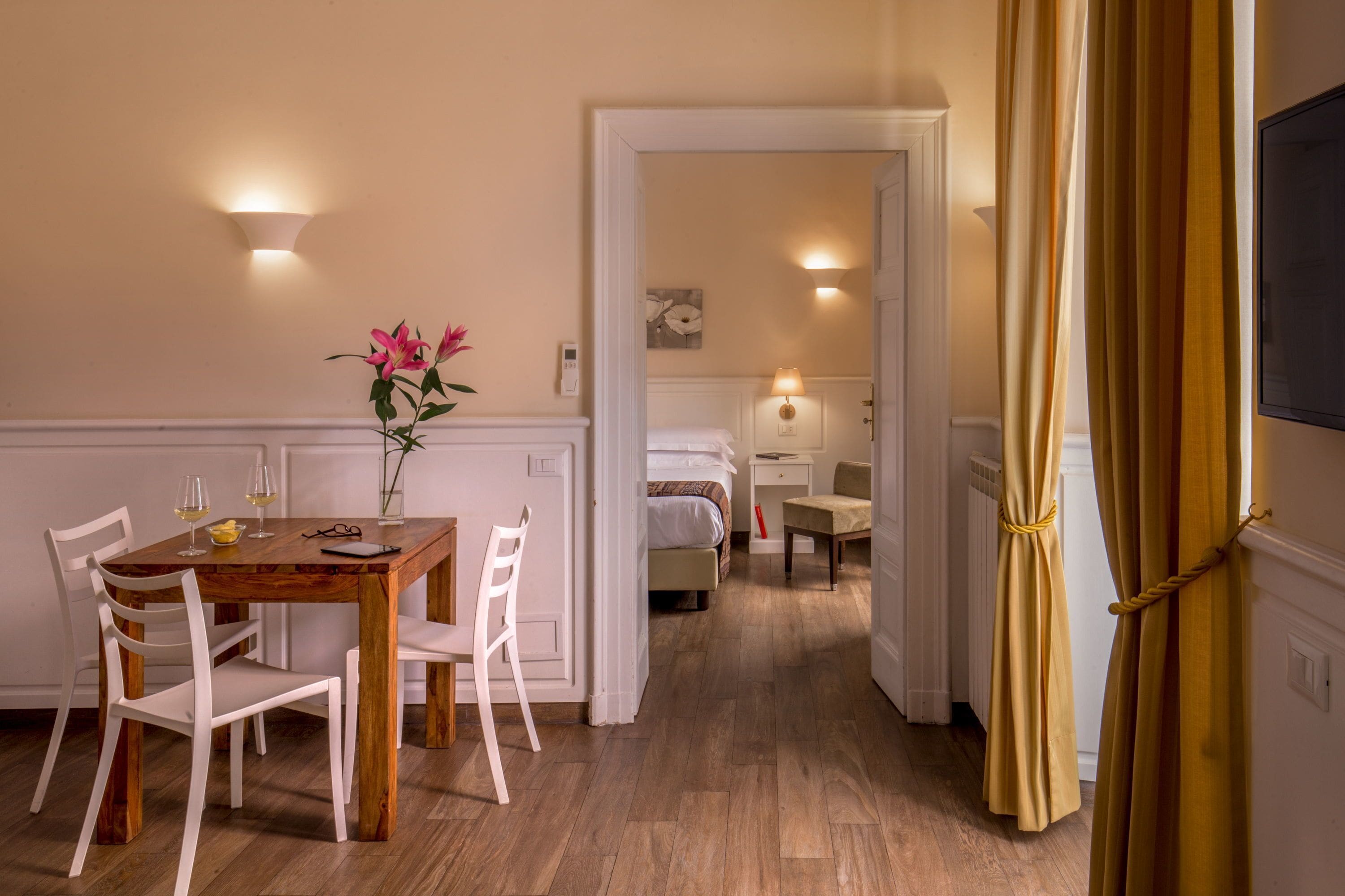 Une chambre d’hôtel avec une table à manger en bois, des chaises blanches, un lit, une table de chevet avec une lampe et une vue sur le couloir.