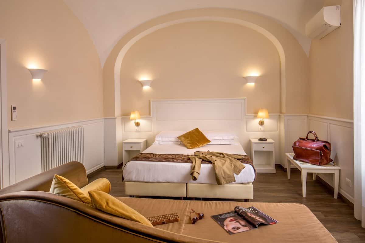 A hotel room with a large bed, two bedside tables, a nightstand, a brown leather sofa, and a magazine on the bed.