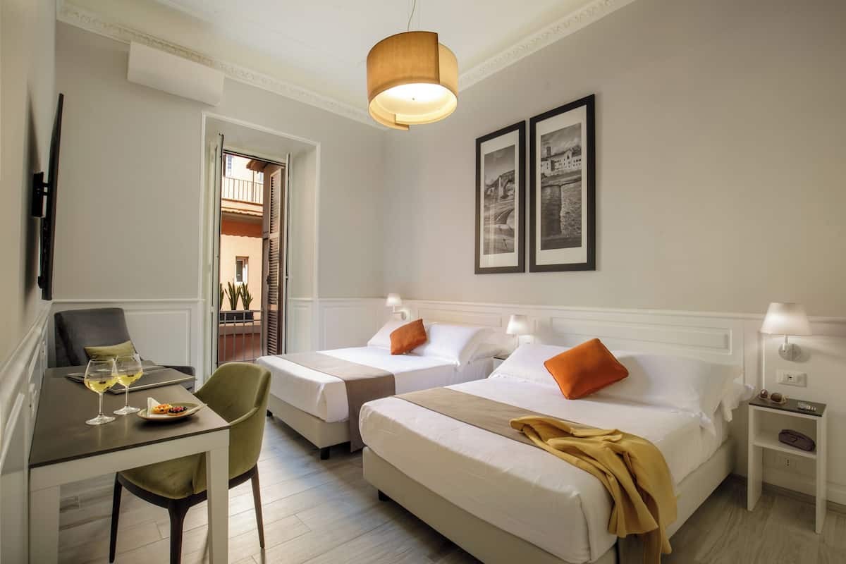 A hotel room with two beds, a dining table with chairs, a small table with a lamp, and framed pictures on the wall.