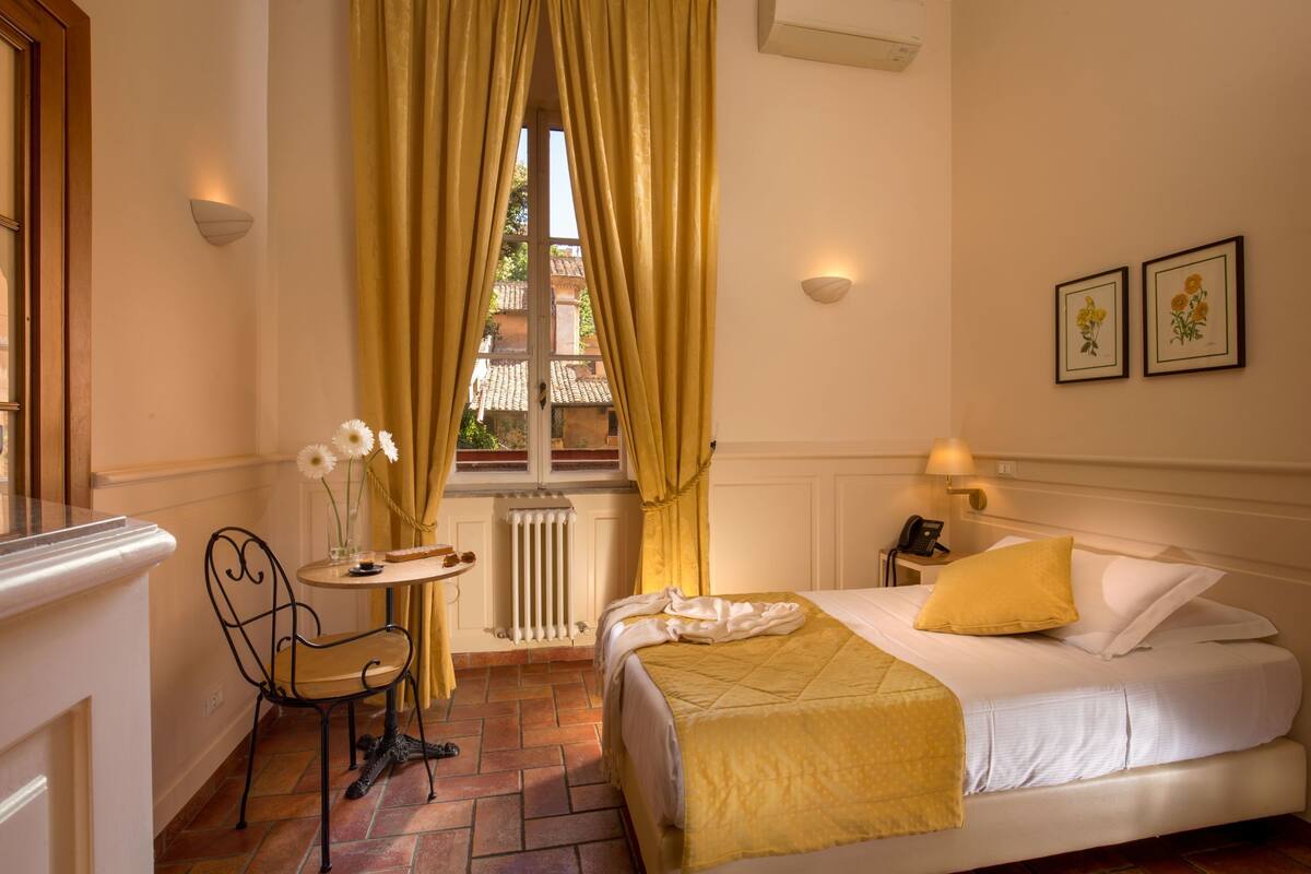 A hotel room with a bed, a chair, a small table with a vase of flowers, and a window with curtains.