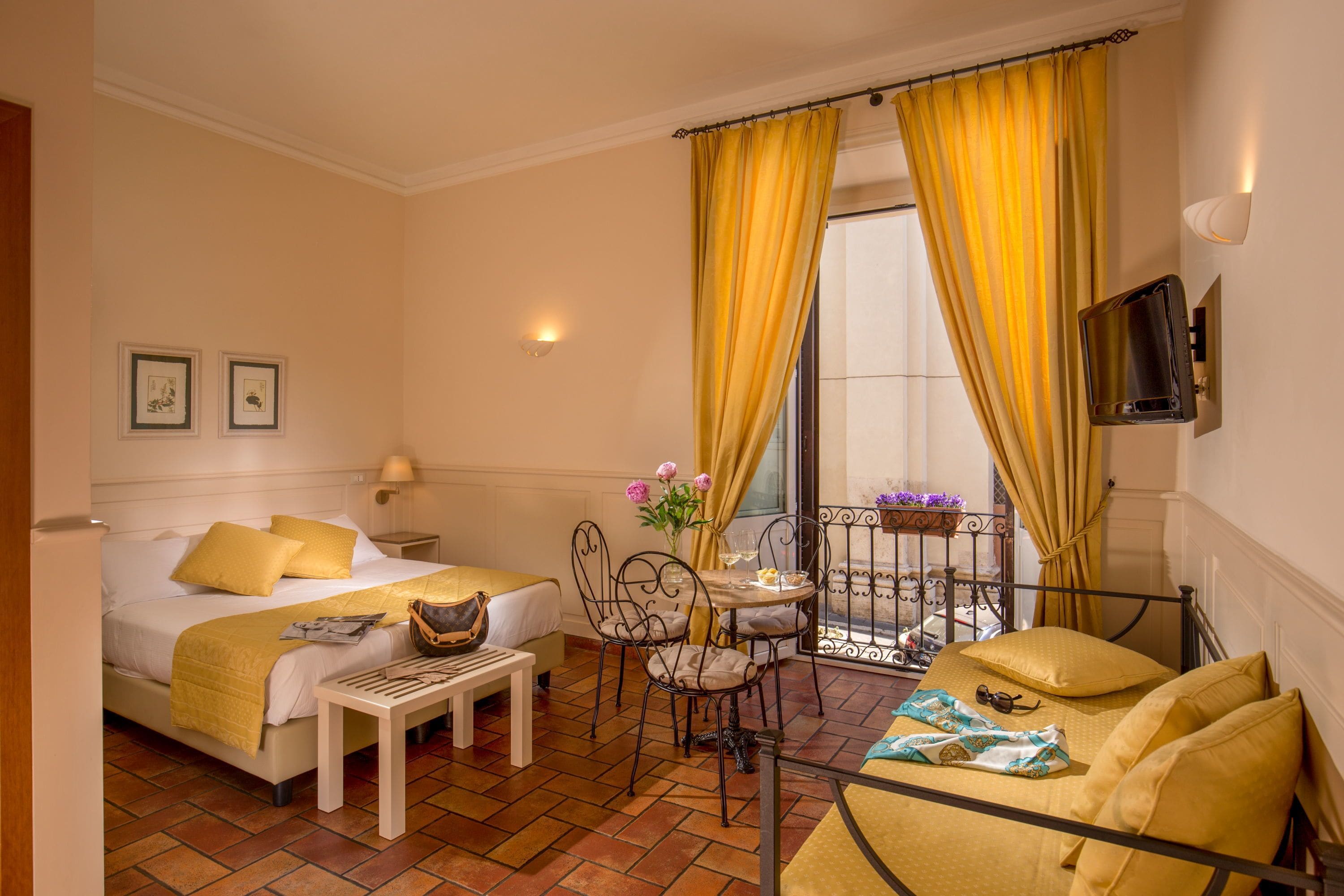 A hotel room with a bed, a desk, a chair, a small table, and a balcony with yellow curtains.