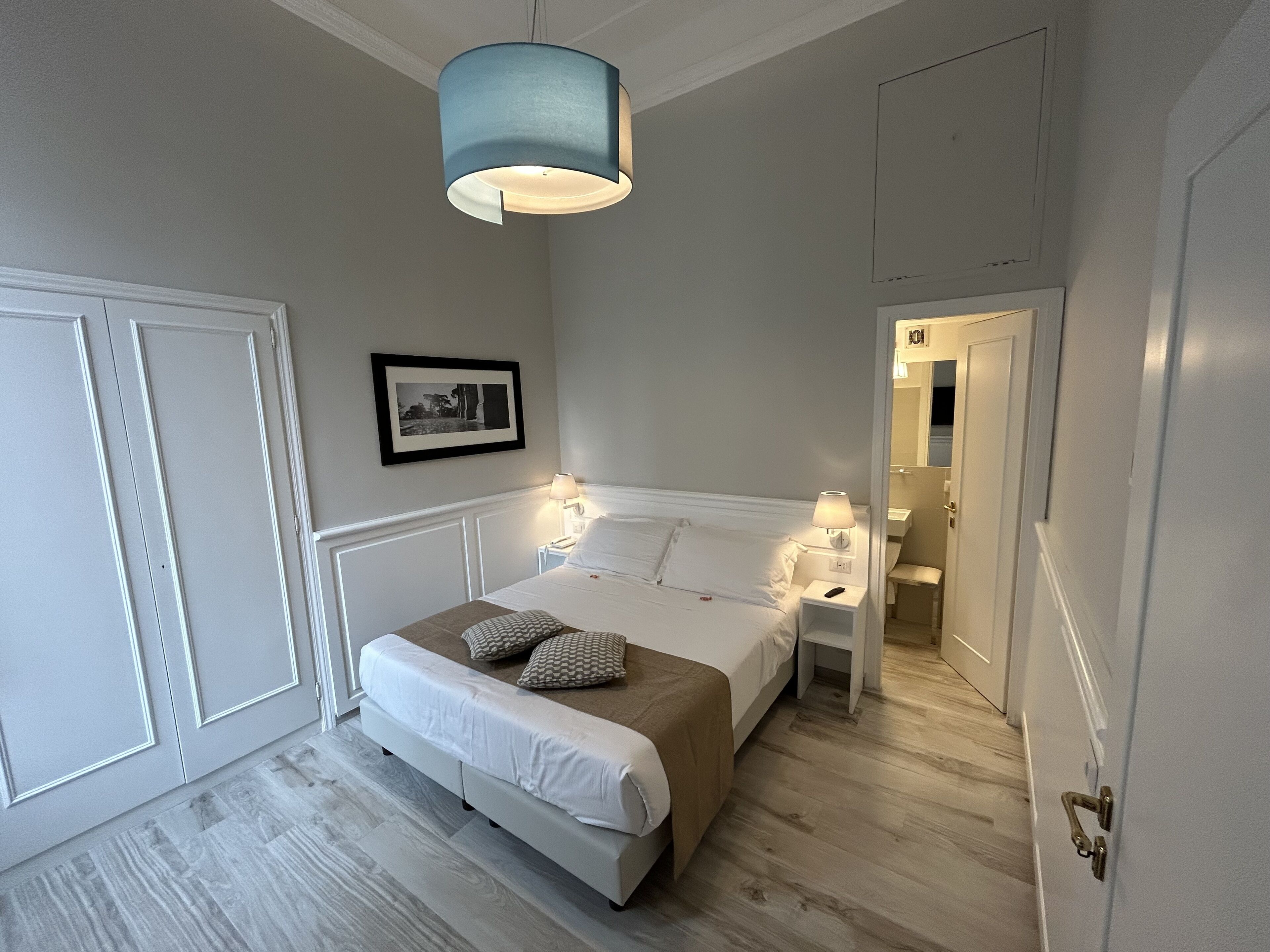 Un dormitorio con cama, mesitas de noche, una lámpara colgante, un cuadro en la pared y una puerta.