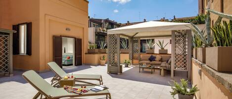 Un patio exterior soleado con sillones, una zona de estar cubierta y plantas en macetas.