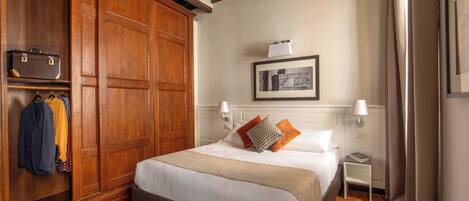 A bedroom with a large wooden wardrobe, a bed with white bedding, a framed picture on the wall, and a small bedside table with a lamp.