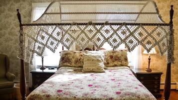 Una cama con dosel y un edredón floral, una mesita de noche de madera con una lámpara, y una silla en una habitación con papel tapiz estampado.